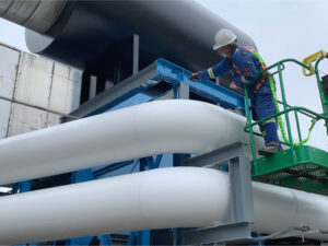 Worker in hard hat and safety harness on a green lift inspecting large white insulated pipes on a blue steel frame.