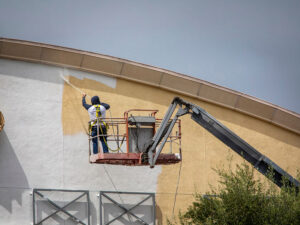 Worker wearing a harness on a raised platform painting the curved beige exterior wall of a building.