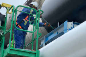 Construction worker in blue coveralls on a green aerial lift painting a blue steel beam beside a large industrial pipe.