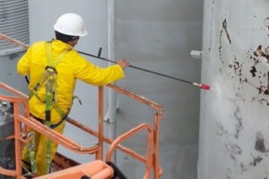 Construction worker in a yellow protective suit and white helmet using a long wand to pressure-wash a concrete wall from an elevated work platform.