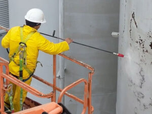 Construction worker in a yellow suit and white helmet stands on an orange scaffold, using a long tool on a concrete wall near the edge of a building surface.