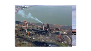 Industrial steel or coal processing plant by a waterfront, with smokestacks emitting white smoke and large piles of material nearby.