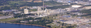 Aerial view of a cement plant with silos, a tall smokestack, conveyors, and pile yards by a river.