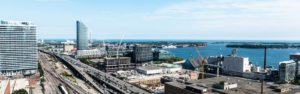 Urban waterfront skyline with a highway and railway tracks cutting through a developing business district by the bay, cranes at construction sites, and modern glass buildings under a blue sky.
