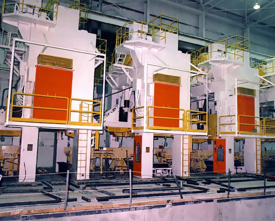 Industrial manufacturing floor with large white machines featuring orange access doors and yellow safety railings and walkways.