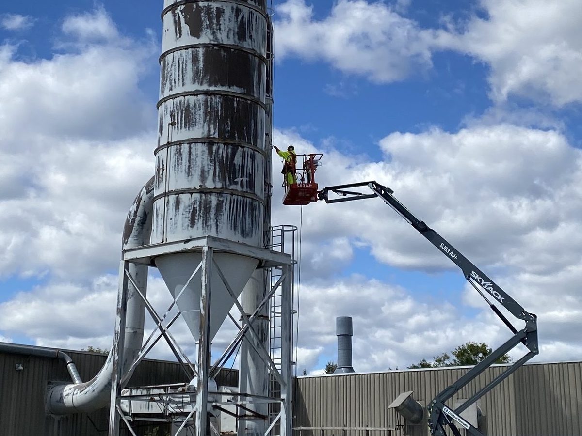 Painter on an aerial lift working on a weathered steel silo at an industrial plant