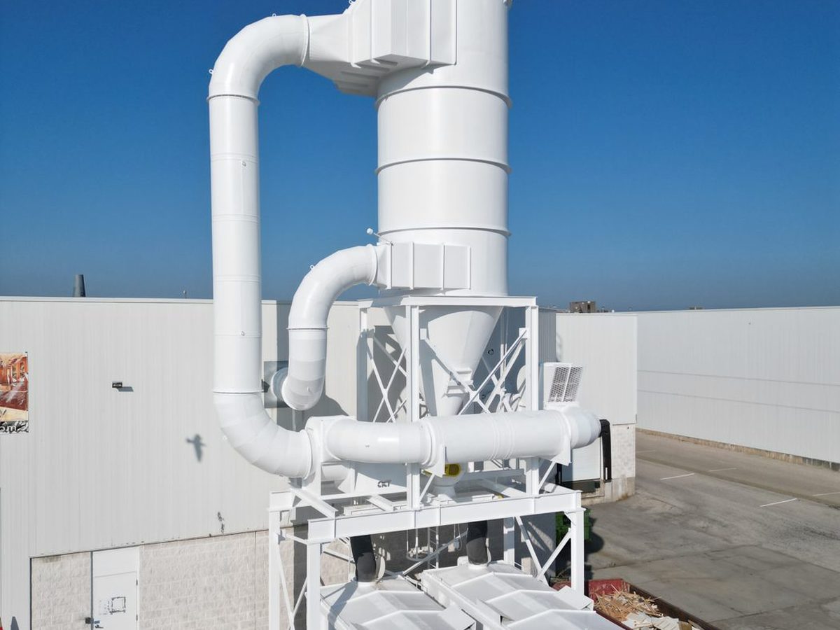 White industrial dust collection tower with ductwork beside a plant building