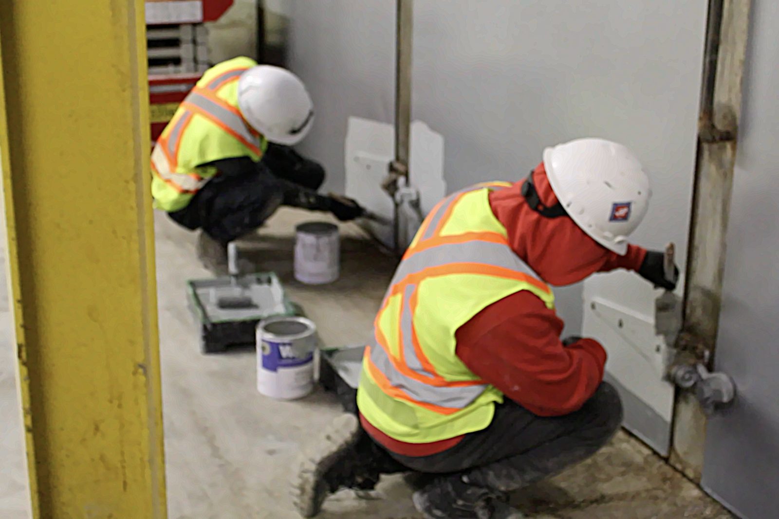 Two painters in high-visibility vests and hard hats painting a wall inside an industrial facility